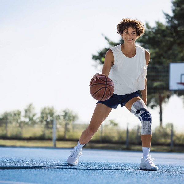 Junge Frau mit weissem Trägershirt, dunkelblauer Sporthose und weisser Kniebandage spielt Basketball auf einem Aussenplatz mit blauem Bodenbelag; sie dribbelt den Ball und lächelt in die Kamera, im Hintergrund ist ein Basketballkorb und Bäume sichtbar.